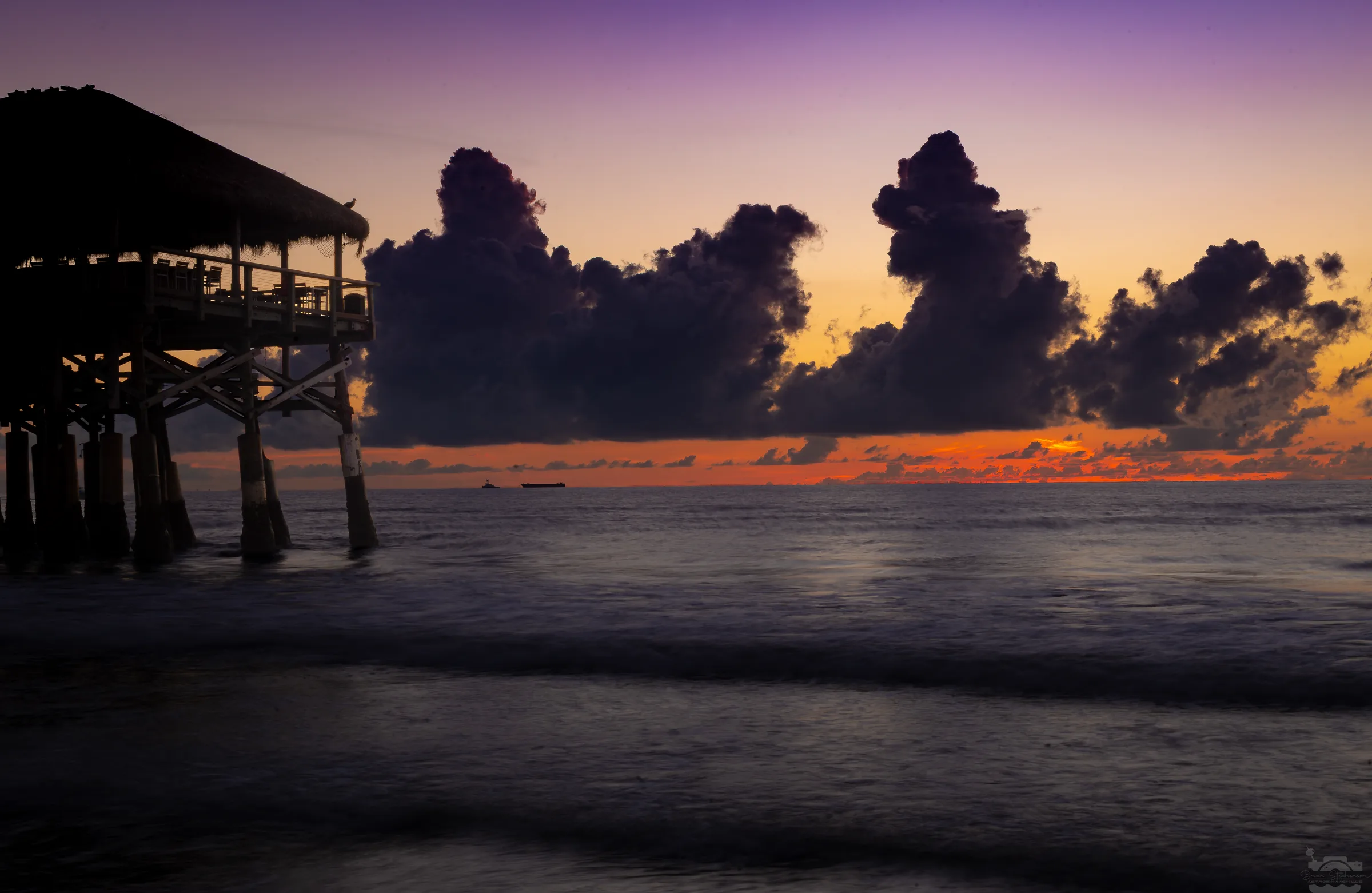 Silhouetted thatched pier against sunrise sky with clouds and calm ocean.